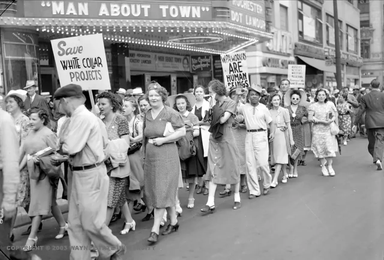 1930s | Large group of primarily women, some carrying picket signs march in front of movie theater, one sign reads, WPA workers are human beings."