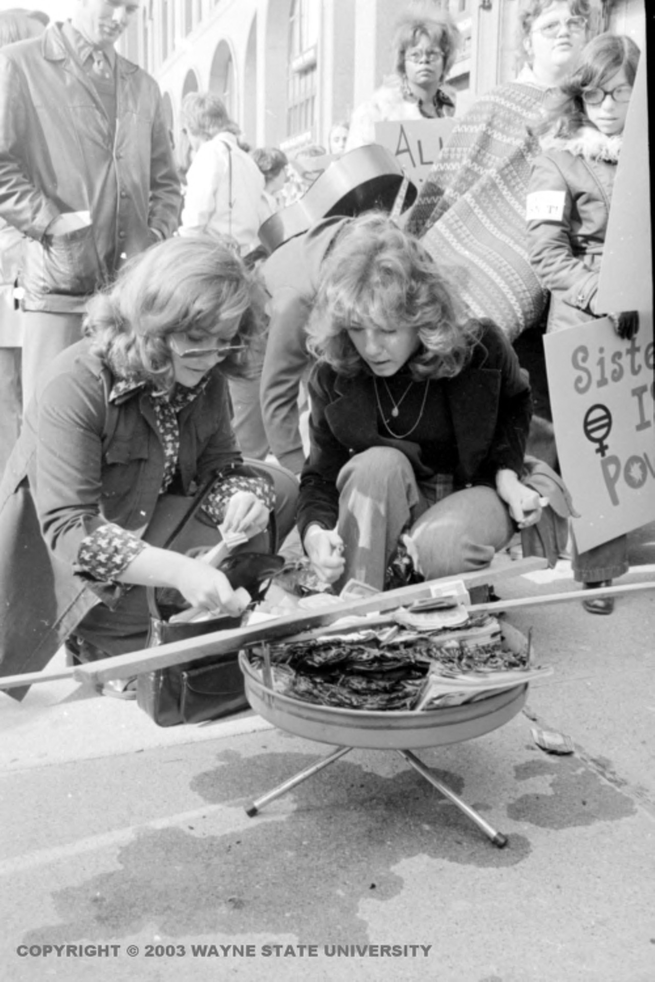 1970s | Two women attempt to light magazines and other materials on fire inside hibachi, at a one day work stoppage organized by the National Organization for Women called Alice Doesn't Day, to draw attention to the work that women perform in society, with women carrying signs behind them.