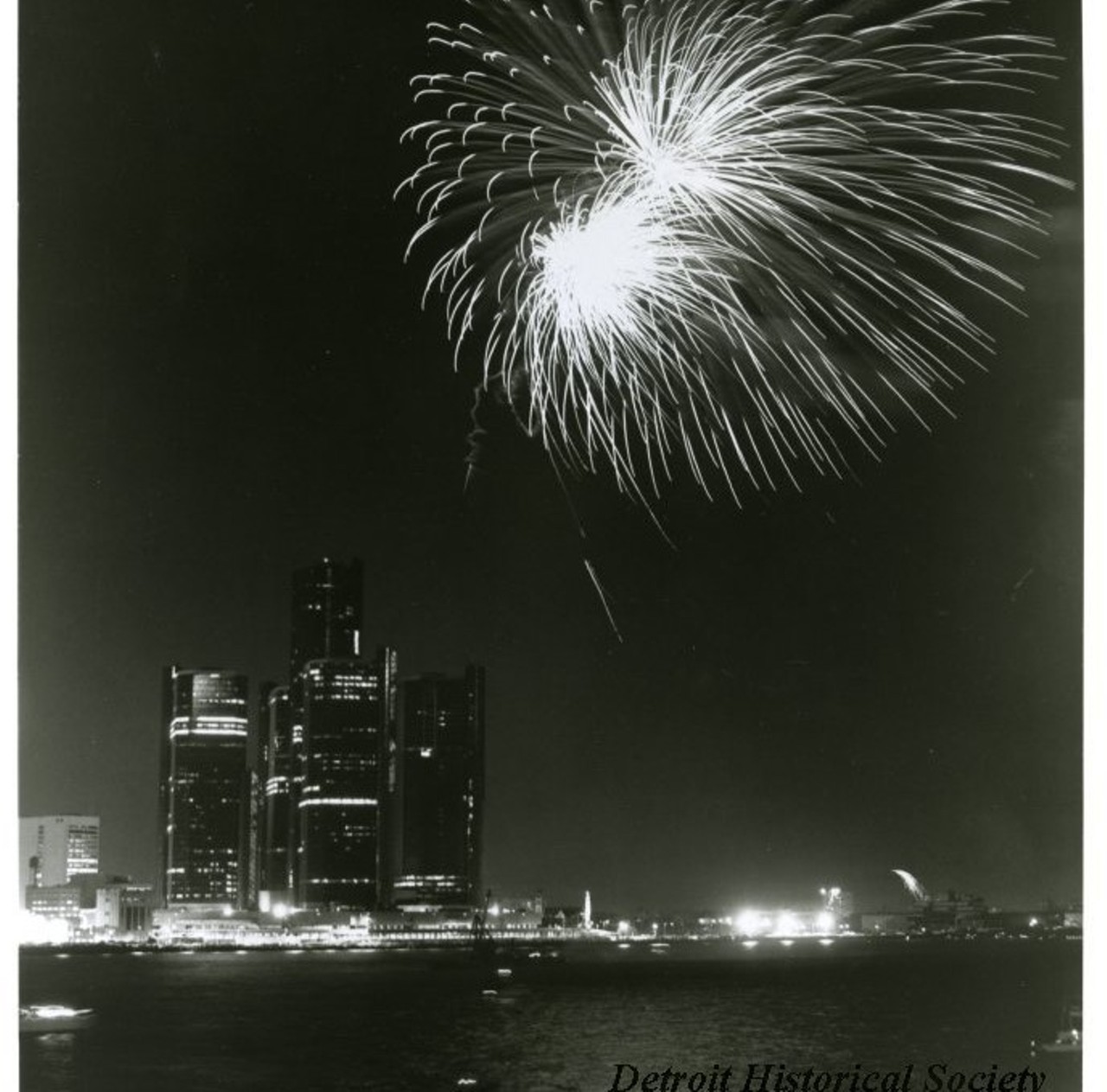 "Black and white photographic print depicting a fireworks display over the Detroit River at night, with the Renaissance Center and Blue Cross Blue Shield Building in the background.
Caption #1 reads- The Hudson's-Stroh's fireworks extravaganza will again be the "crowning jewel" in the events planned for the Detroit-Windsor International Freedom Festival, June 30 - July 4. This year's fireworks display will begin downtown at dusk on June 30 and will conclude after several tons of colorful starbursts have been shot into the night sky from barges on the Detroit River. In case of bad weather, everything will be rescheduled for July 1. The fireworks is only one of over 75 Festival events.
Caption #2 reads- For release on or after Tuesday, June 5, 1979. Detroit River "lights up" June 29 - For the 21st consecutive year, the spectacular Freedom Festival Fireworks will herald the opening of Detroit and Windsor's annual celebration. This year's fireworks display will begin at 9:55 p.m., Friday, June 29, from barges anchored midway in the Detroit River opposite the Veterans Memorial Building, Cobo Hall and Ford Auditorium. More than 600,000 people are expected to watch the seven tons of colorful starbursts light up the skylines of the two cities. In case of bad weather, everything will be rescheduled for Saturday, June 30."