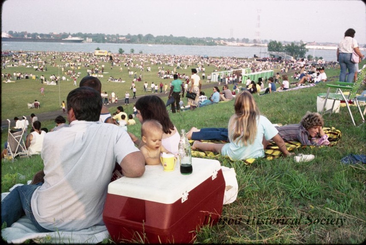 "Color negative taken of a crowd gathered on the parade grounds of Fort Wayne, likely to watch the Windsor–Detroit International Freedom Festival fireworks display, as taken from the north end of the hill at the west side of the field. In the foreground a baby leans on a red cooler between two adults. Other groups of spectators seated on blankets or in lawn chairs spot the grounds. A bank of portable toilets have been installed along the path at the foot of the hill. A stage has been set up along the riverfront. A freighter is visible on the Detroit River in the background. The film was previously housed in a GK Photographics envelope, with "AAA 4-20-88, Fort Wayne," handwritten on it."