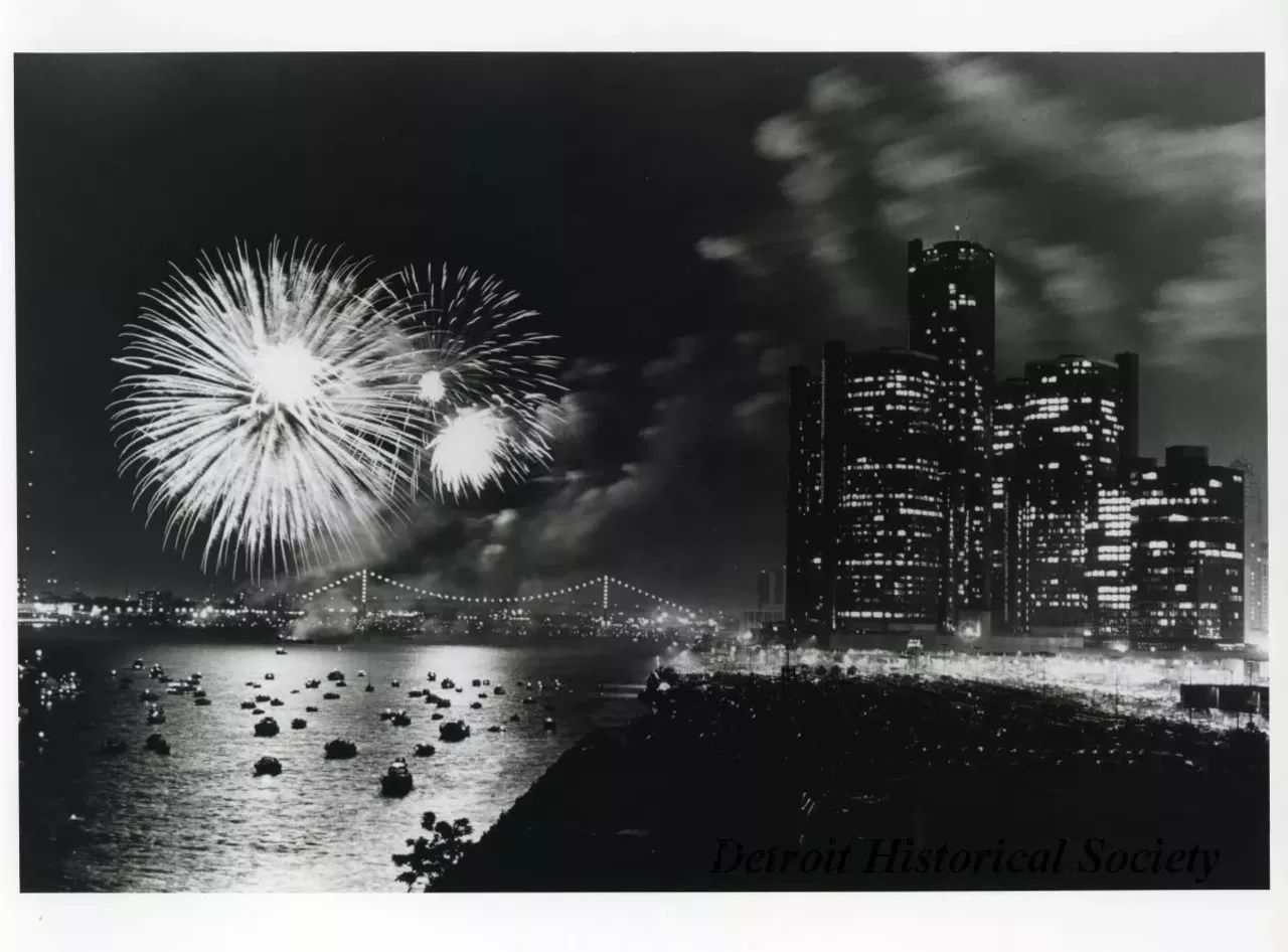 "Black and white photographic print depicting a fireworks display over the Detroit River at night, with the Ambassador Bridge to the left and the Renaissance Center to the right. The river is filled with small boats. Caption reads- Freedom Festival Fireworks - One of the highlights of the 1984 Detroit-Windsor International Freedom Festival was the riverfront fireworks display captured in this photograph by Mark Cornillie. It appeared in newspapers around the country. An estimated one million people turn out for the show sponsored by Hudson's, Stroh's, WDIV-TV 4 and WMJC-FM. Handwritten on verso: Used June 1985 Michigan Living"