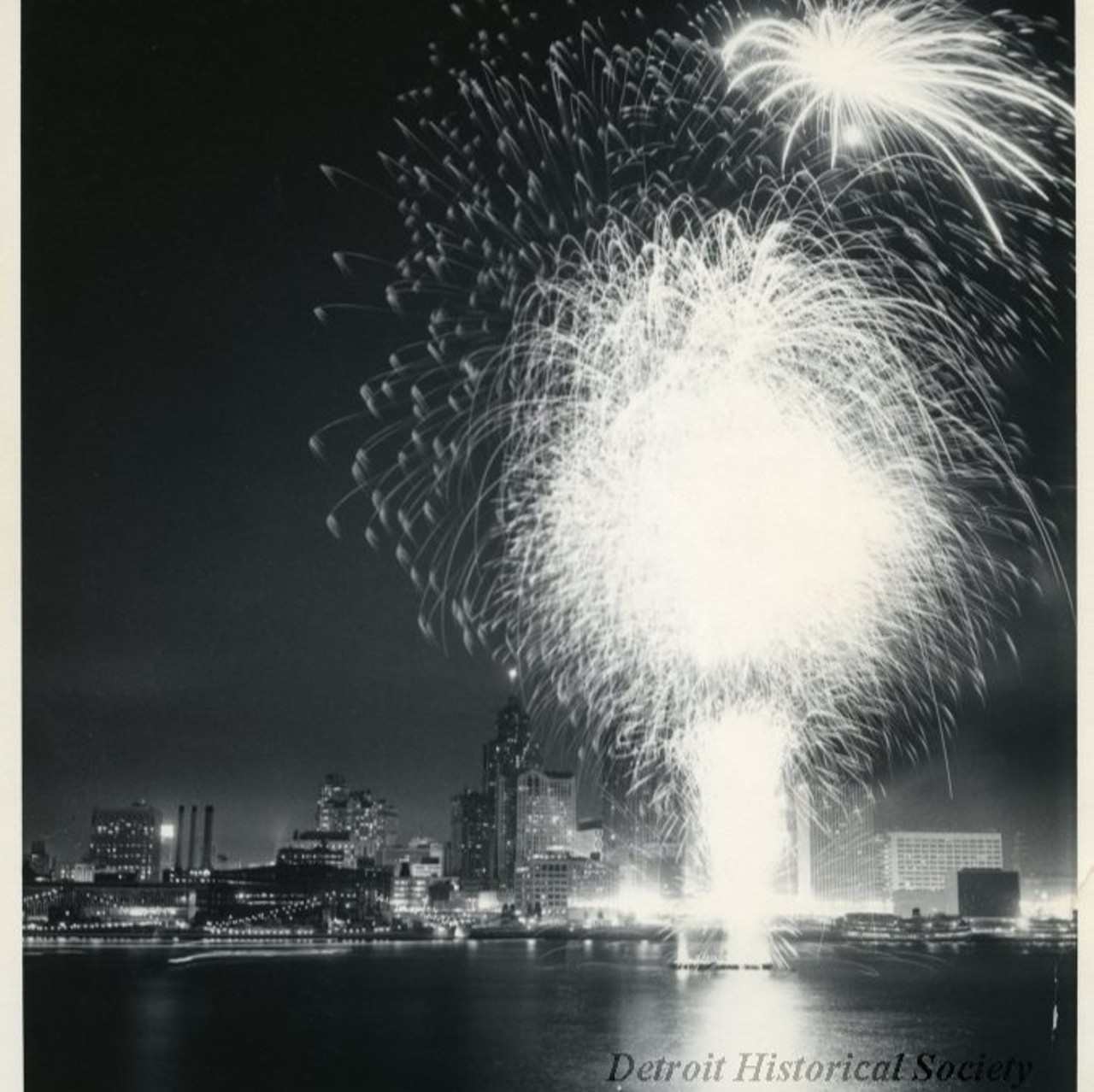 "Black and white photographic print depicting a fireworks display over the Detroit River at night, with the downtown skyline in the background. In view are Ford Auditorium, the City-County Building, Buhl Building, and Cobo Arena under construction.
Caption reads- Freedom is a Happy Thing: The largest crowd in the history of Detroit and Windsor ever to assemble for a single event, an estimated million people, saw the J.L. Hudson sponsored fireworks, highlight of the annual International Freedom Festival (Detroit-Windsor). The spectacular fireworks took place off barges in the middle of the Detroit River.
Handwritten on verso: Used in Motor News June, 1960"