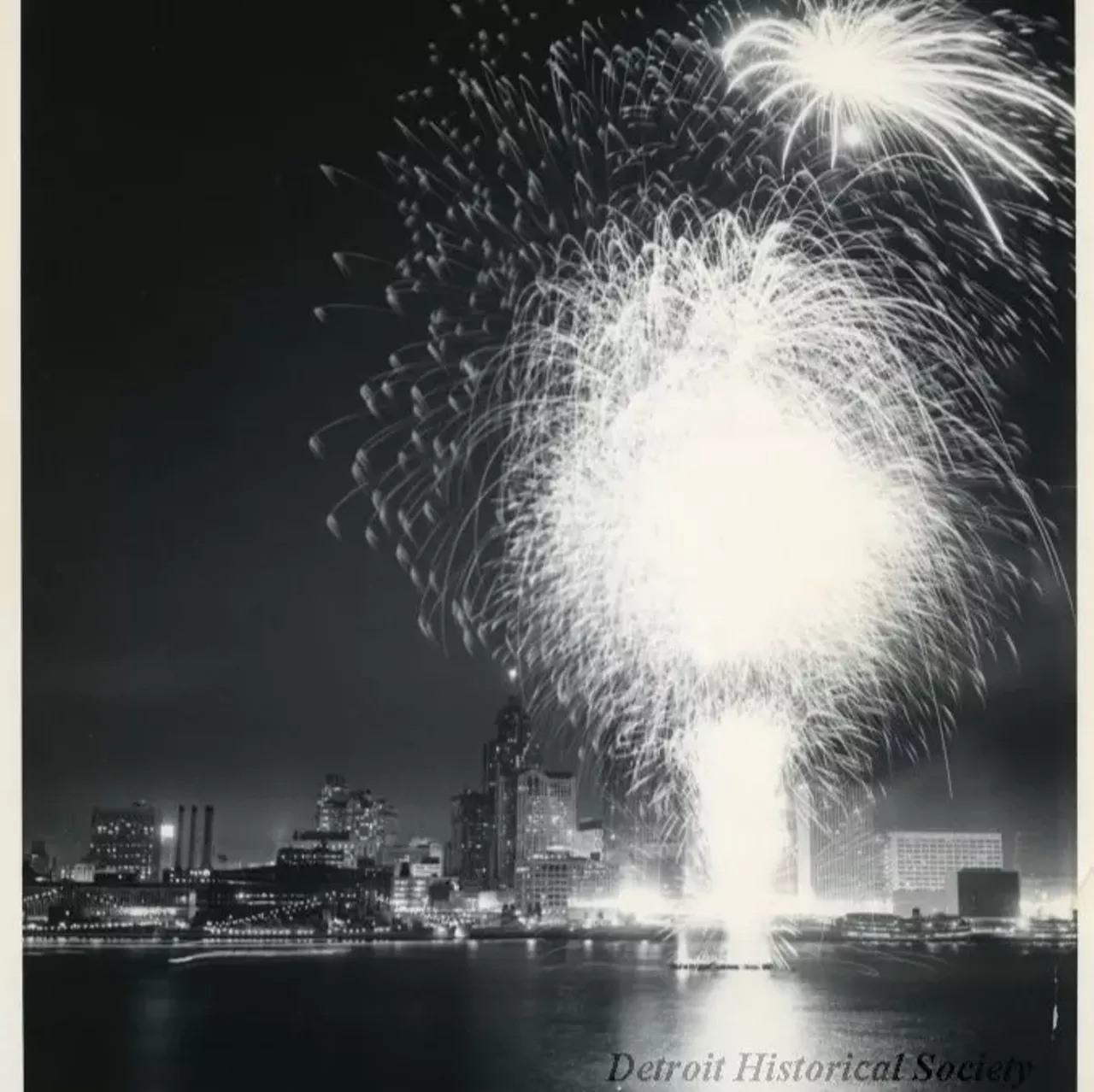 "Black and white photographic print depicting a fireworks display over the Detroit River at night, with the downtown skyline in the background. In view are Ford Auditorium, the City-County Building, Buhl Building, and Cobo Arena under construction. Caption reads- Freedom is a Happy Thing: The largest crowd in the history of Detroit and Windsor ever to assemble for a single event, an estimated million people, saw the J.L. Hudson sponsored fireworks, highlight of the annual International Freedom Festival (Detroit-Windsor). The spectacular fireworks took place off barges in the middle of the Detroit River. Handwritten on verso: Used in Motor News June, 1960"