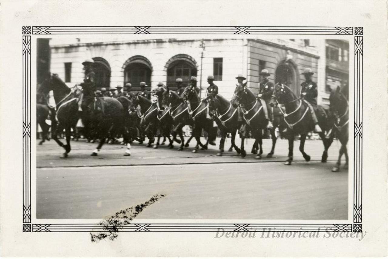 "Black and white photographic print with a decorative border taken at the 1932 Fourth of July parade on Michigan Avenue at the intersection of Griswold Street facing northeast. A group of mounted police officers ride in two lines along Michigan. The Peoples Wayne County Bank stands in the background. "Jul 4 1932," and "108," are stamped on the verso."