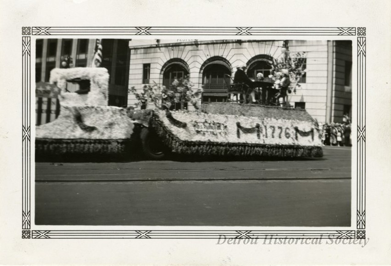 "Black and white photographic print with a decorative border taken at the 1932 Fourth of July parade on Michigan Avenue at Griswold Street facing northeast. A parade float consisting of a flower-covered car pulling a similarly decorated trailer atop of which a group of men in 18th century costume sit around a table. A sign reading, "U.S. Rubber Company," is attached to the side of the trailer, next to where "1776" is written in flowers. A crowd stands in the background along Michigan in front of the Peoples Wayne County Bank. "Jul 4 1932," and "108," are stamped on the verso."