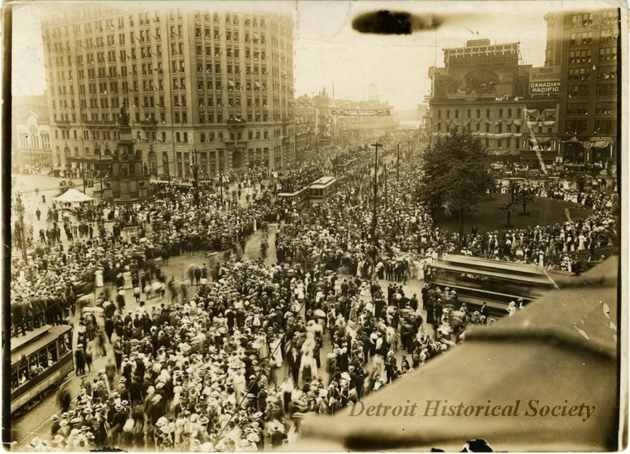 "Sepia-toned photographic print from the studio of Louis James Pesha depicting a large crowd gathered in Campus Martius, for the automobile parade held as part of the Cadillaqua celebration overlapping with the Fourth of July, as viewed from an elevated position, likely at the Majestic Building, facing south. A banner, reading, "Harold Jarvis will sing of Masonic Moonlight on steamer STE. CLAIRE," "City of the Straits Lodge, Mon. July 29," and "Mon. July 29."
The majority of the crowd are wearing hats, and boater hats appear to be the predominant style. Streetcars make their way through the crowd, and those headed north on Woodward Avenue carry additional passengers on their roofs. A utility pole near the Michigan Soldiers' and Sailors' Monument has become a perch for several spectators as well. American flags are hung from nearly every other window of the Pontchartrain Hotel. A large sign reading, "Conventions & Auto Bulletin" is posted above the corner of Woodward and Fort Street. To its right are several signs advertising railroad lines—Canadian Pacific, Pere Marquette, Union Pacific and Southern Pacific. This corner is also draped with flags and banners. The stretch of Woodward leading to the Detroit River is similarly decorated. "Detroit, Mich. 1907, NA 66537-B" handwritten on verso."