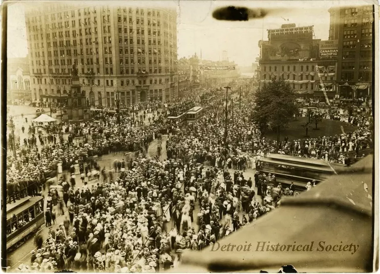 "Sepia-toned photographic print from the studio of Louis James Pesha depicting a large crowd gathered in Campus Martius, for the automobile parade held as part of the Cadillaqua celebration overlapping with the Fourth of July, as viewed from an elevated position, likely at the Majestic Building, facing south. A banner, reading, "Harold Jarvis will sing of Masonic Moonlight on steamer STE. CLAIRE," "City of the Straits Lodge, Mon. July 29," and "Mon. July 29." The majority of the crowd are wearing hats, and boater hats appear to be the predominant style. Streetcars make their way through the crowd, and those headed north on Woodward Avenue carry additional passengers on their roofs. A utility pole near the Michigan Soldiers' and Sailors' Monument has become a perch for several spectators as well. American flags are hung from nearly every other window of the Pontchartrain Hotel. A large sign reading, "Conventions & Auto Bulletin" is posted above the corner of Woodward and Fort Street. To its right are several signs advertising railroad lines—Canadian Pacific, Pere Marquette, Union Pacific and Southern Pacific. This corner is also draped with flags and banners. The stretch of Woodward leading to the Detroit River is similarly decorated. "Detroit, Mich. 1907, NA 66537-B" handwritten on verso."