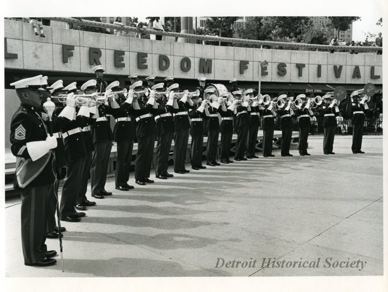 "Black and white photographic print depicting a military brass band performing in the Hart Plaza amphitheater In the background is the Down Under restaurant and people watching from above.
Caption reads: Military bands have traditionally been a highlight of the International Freedom Festival. This year the Festival will showcase the U.S.A.F. Presidential Drill Team, Tops in Blue, the First Marine Band and other musical groups representing the talents of our armed forces."