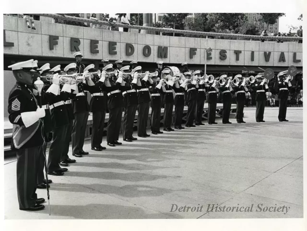 "Black and white photographic print depicting a military brass band performing in the Hart Plaza amphitheater In the background is the Down Under restaurant and people watching from above. Caption reads: Military bands have traditionally been a highlight of the International Freedom Festival. This year the Festival will showcase the U.S.A.F. Presidential Drill Team, Tops in Blue, the First Marine Band and other musical groups representing the talents of our armed forces."