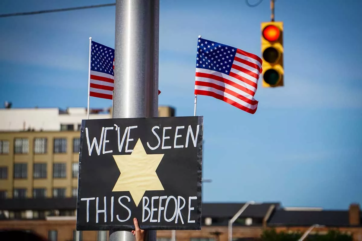 Image: A sign with American flags reading, “We’ve seen this before” with a Jewish yellow star, held at a protest in Detroit in 2018.