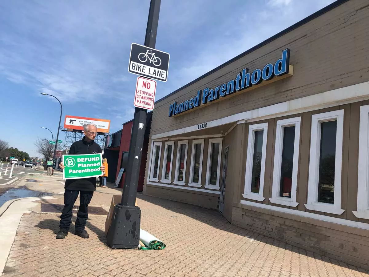 Image: A protester stands outside a Ferndale Planned Parenthood clinic holding a sign that says “Defund Planned Parenthood.”