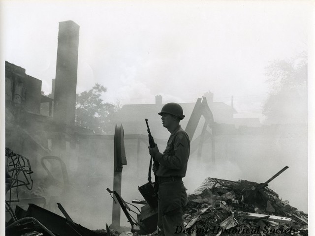 "One black and white photograph of a Michigan National Guardsman who is on patrol at the corner of 12th and Hazelwood Streets in the aftermath of the 1967 Detroit Riot. The smoldering ruins of several buildings are visible in the background."
