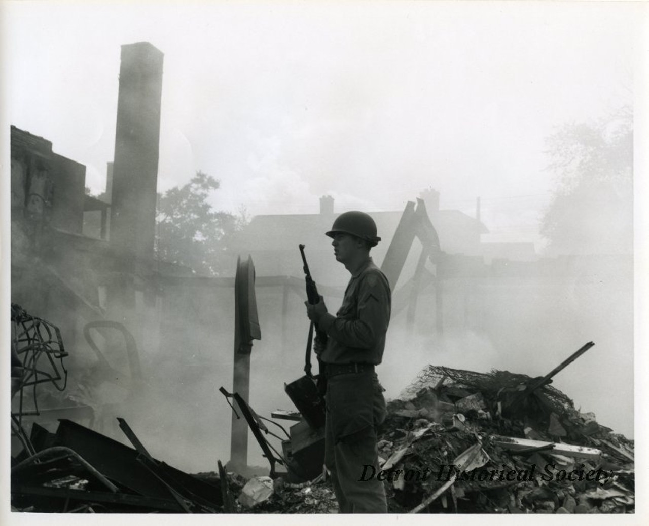 "One black and white photograph of a Michigan National Guardsman who is on patrol at the corner of 12th and Hazelwood Streets in the aftermath of the 1967 Detroit Riot. The smoldering ruins of several buildings are visible in the background."