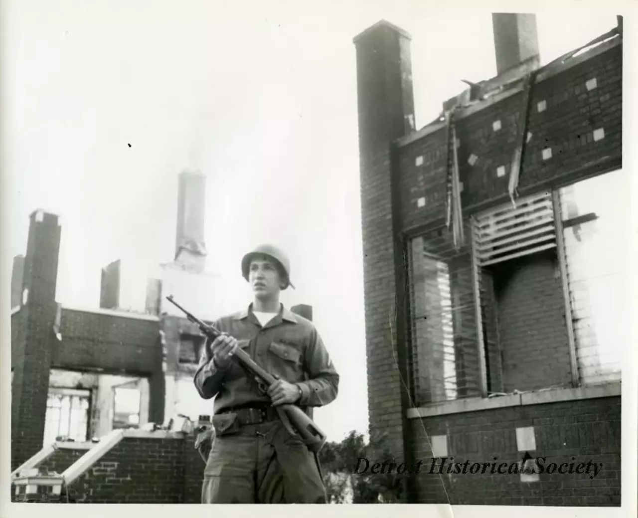 "One black and white photograph of a Michigan National Guardsman who is on patrol in the aftermath of the 1967 Detroit Riot. The ruins of several 2-story brick buildings are visible in the background."