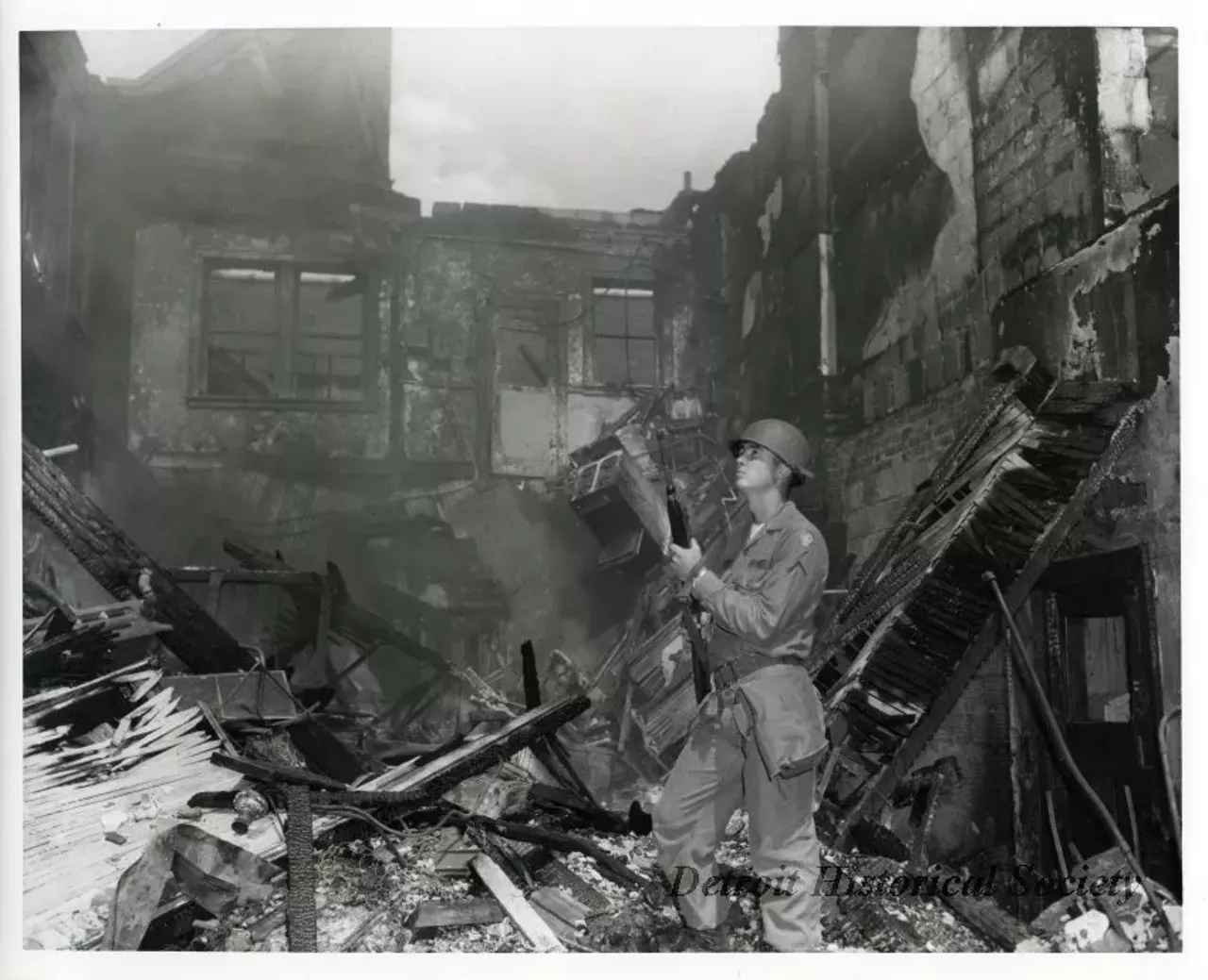 "One black and white photograph of a Michigan National Guardsman who is on patrol in the aftermath of the 1967 Detroit Riot. The smoldering ruins of a partially collapsed 2-story building are visible in the background."