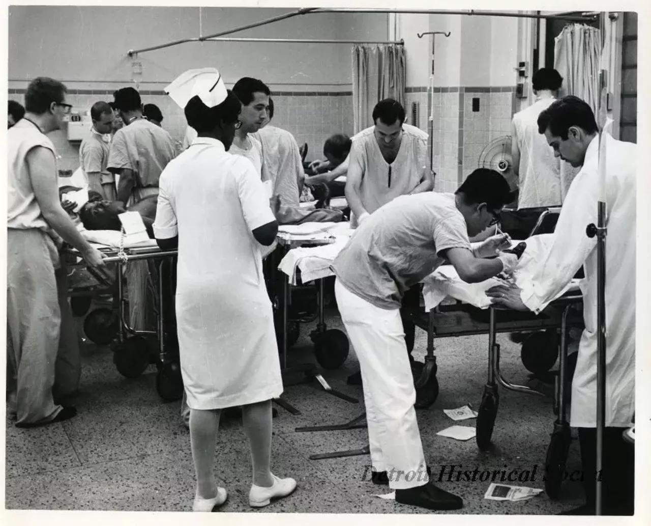 "One black and white photograph of the hospital emergency room scene (probably Detroit Receiving Hospital) after the arrival of injury victims from the 1967 Detroit Riot. Doctors, nurses, and other medical personnel are attending to injured persons who are laid out on gurneys."