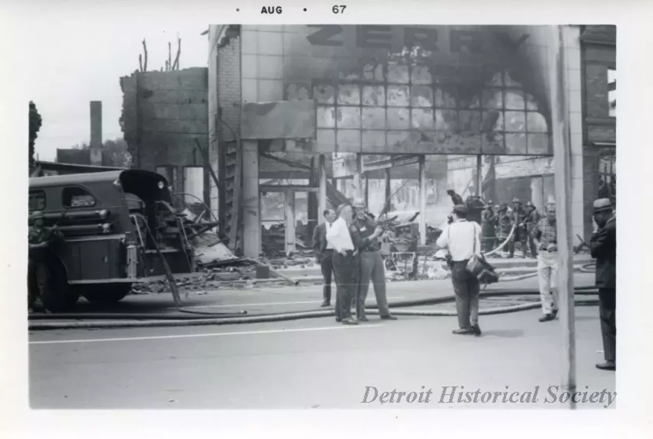 "One digital scan of a black and white photograph which shows several stores that have been destroyed by fire during the 1967 Detroit Riot. The image shows Governor George Romney (in white shirt) in the center foreground. He is standing on the side of a street and is speaking with a Michigan State policeman. A news photographer and several other persons are standing nearby on the street. A Detroit Fire Department truck, firemen, and the ruins of the Zerry Furniture store can be seen in the background."