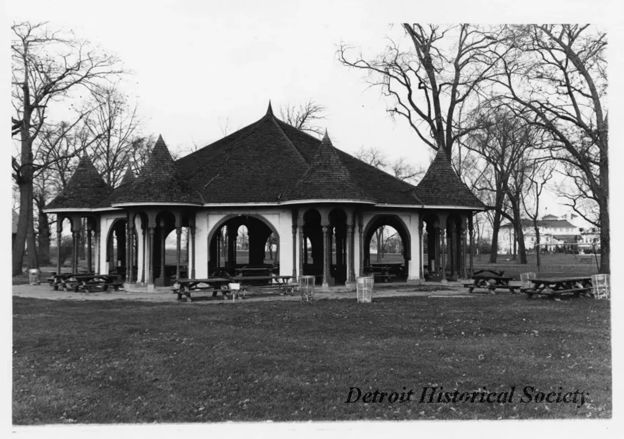"Black and white photographic print depicting a picnic shelter on Belle Isle, with picnic tables and trash cans in front. In the background is the Detroit Boat Club. Dated December, 1973."