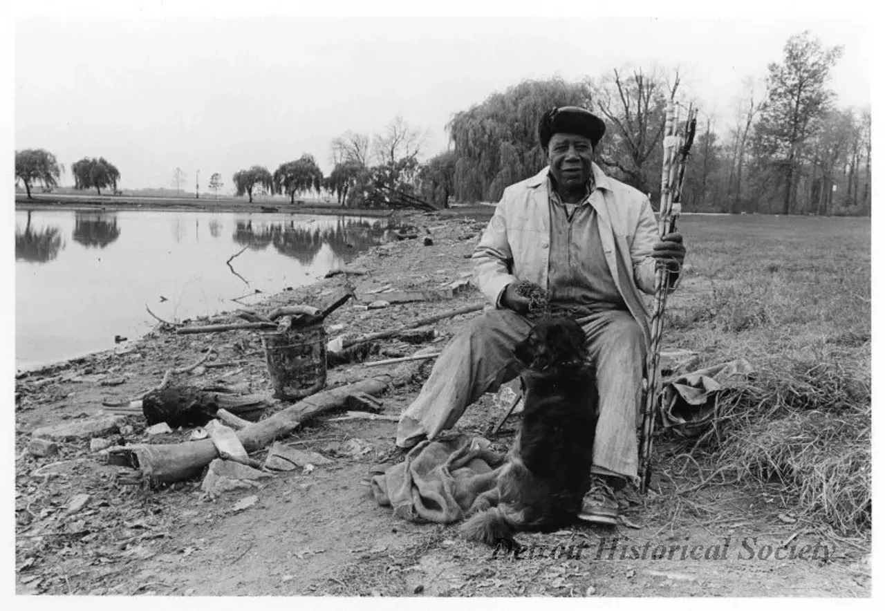 "Black and white photographic print depicting a seated African American man with a dog sitting at his feet near a lake on Belle Isle. The man is holding the dog's chain with one hand and fishing rods with the other. A metal bucket with pieces of wood is visible. Dated November 1973."