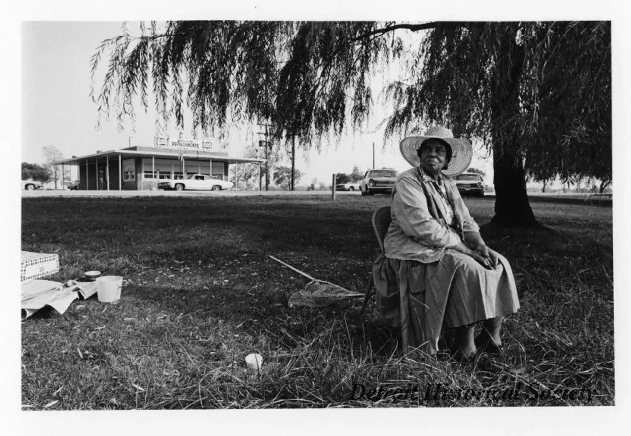 "Black and white photographic print mounted on board depicting older African-American woman sitting in a metal folding chair by a willow tree on Belle Isle. A large fishing net, fishing rods on newspaper and a plastic bucket lay in the grass around her. A concession stand and parked cars are visible in the background. A sign on the concession stand reads 'Pepsi Refreshments'."