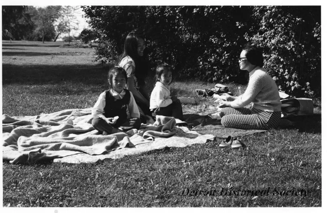"Black and white photographic print mounted on board depicting two Asian women and young girls sitting on blankets in the grass on Belle Isle. Dated October 1973."
