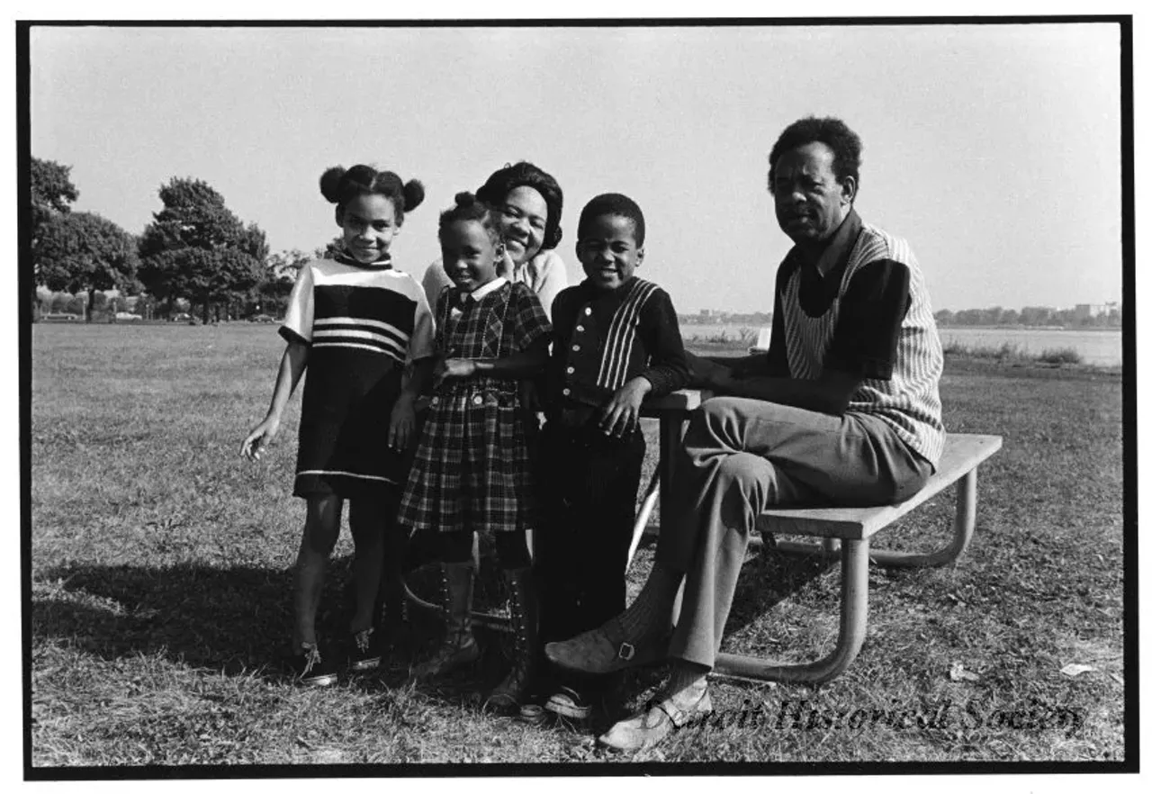  "Black and white photographic print mounted on board depicting an African American family of five at a picnic table on Belle Isle. Parked cars and the Detroit River are visible in the background." Circa 1973.