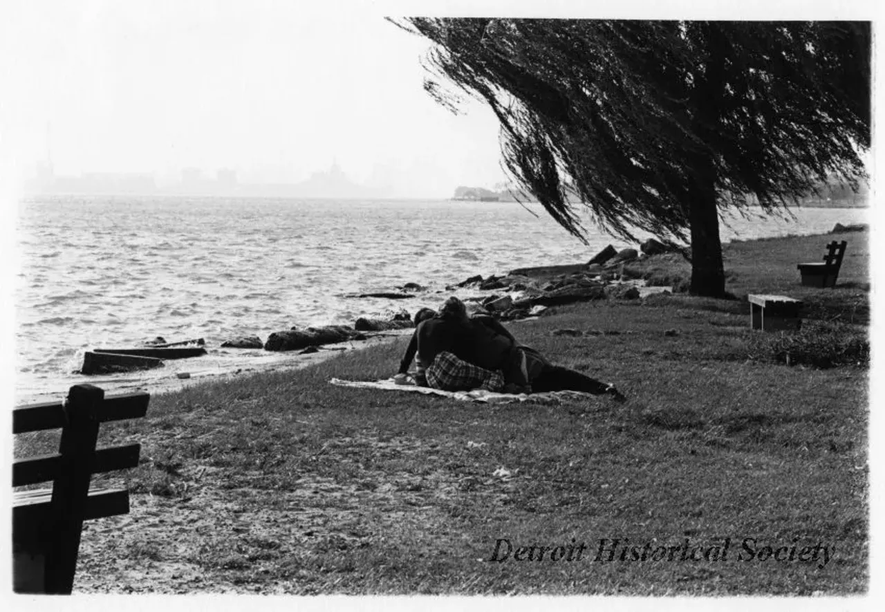 "Black and white photographic print mounted on board depicting a couple lying on a blanket by the shore on Belle Isle. Several benches visible in picture." Circa 1973.