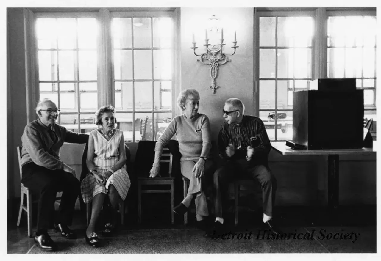 "Black and white photographic print on board depicting four senior citizens seated in chairs inside the Belle Isle Casino. A sconce with four candles hangs on the wall just above them. Dated November 1973."