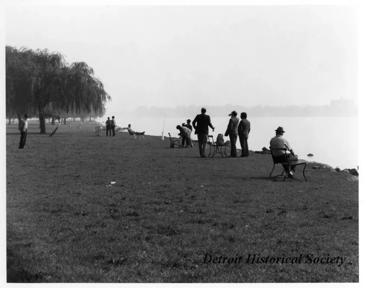 "Black and white photographic print mounted on board depicting people gathered at the benches on the Belle Isle shore. Fishing poles are propped up along the edge of the shore."