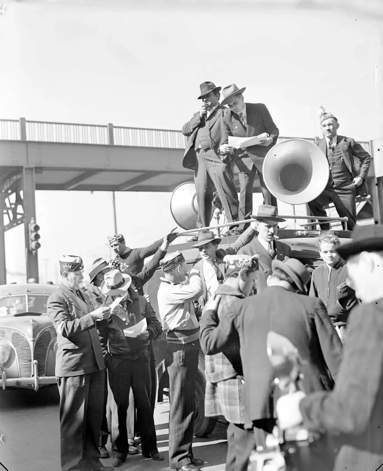  April 11, 1941: Strike ends! A man stands on top of an automobile and speaks through loudspeakers as a group of men stand on the ground near the car, some wearing UAW hats.